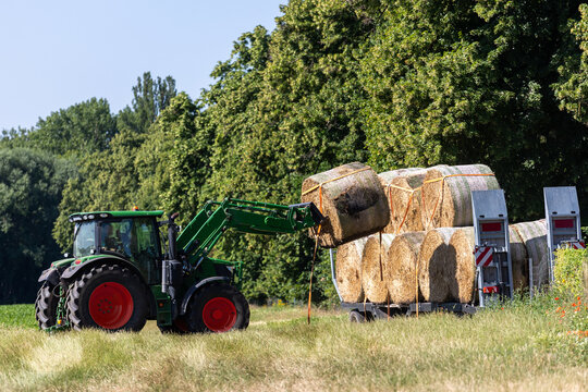 Green tractor loads round hay bale onto farm trailer stacked with straw beside meadow. Summer harvesting scene shows rural machinery, agriculture, transport, fieldwork, and sunny countryside