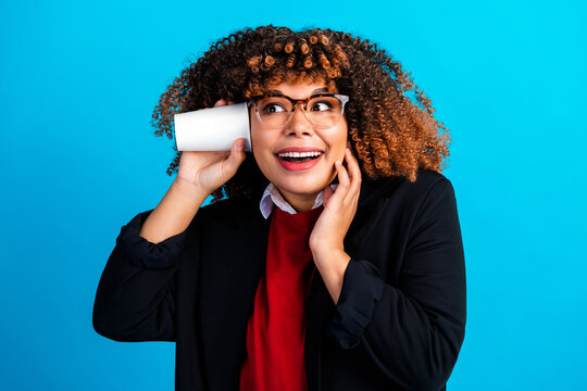 Young woman listening with a paper cup phone, playful communication in business casual with glasses and curly hair against a bright blue background
