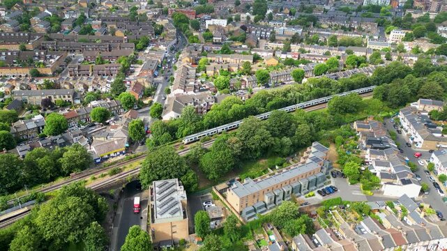 Aerial view of a train passing through the residential neighborhood of Peckham, featuring rows of brick houses, lush green trees, and urban infrastructure in London, England, United Kingdom.