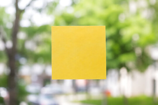 Yellow sticky note on glass window, blank adhesive paper for reminders, square memo pad note with blurred outdoor park background isolated on White Background ai