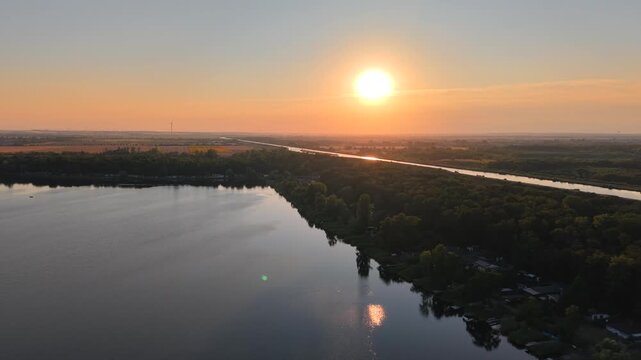 Drone view captures lakeside holiday settlement, sailboats, and Mittelland Canal near Magdeburg at sunset. Golden light spreads across woodland, calm water, gardens, and distant rural horizon