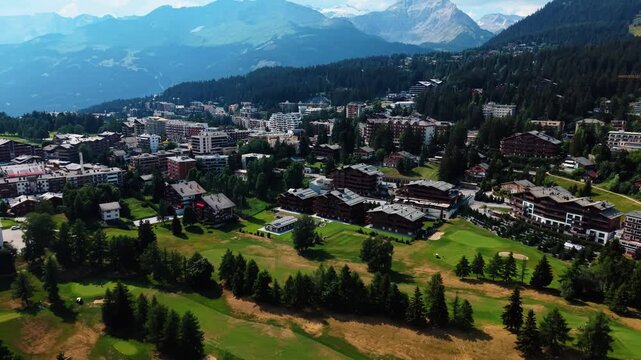 Aerial view of the Guarda Golf Hotel and Residences surrounded by lush green golf courses and alpine architecture in Crans-Montana, Valais, Switzerland.