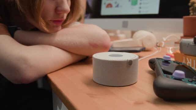 Caucasian teenager resting head on desk while looking at smart speaker displaying time and temperature. Footage is ideal for smart home technology ads and modern youth lifestyle promos