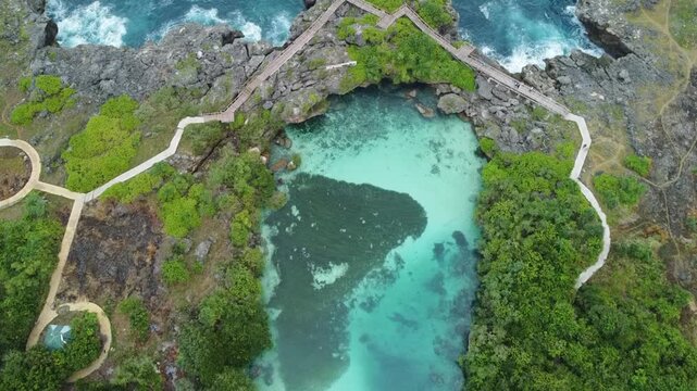 Aerial view of natural turquoise lagoon surrounded by rocky cliffs and lush vegetation with wooden walkways in Sumba, East Nusa Tenggara, Indonesia.