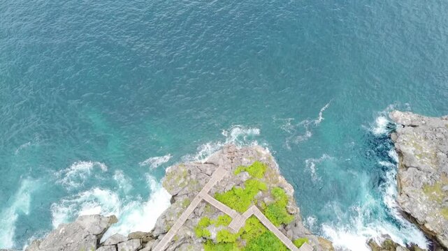 Aerial view of a rocky cliff with a wooden walkway surrounded by turquoise ocean waves in Sumba, Indonesia.