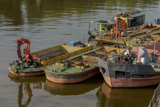 Industrial barges with crane manipulators on river water surface near embankment. Cargo vessels and workboats for technical riverbed cleaning and shipping maintenance.