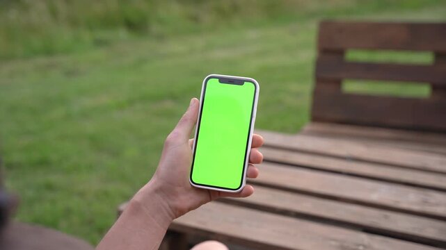 Hand holds phone with green screen outdoors. Person sits on wooden bench near grass. Phone display shows placeholder for content. Suitable for apps, marketing, or UI mockups
