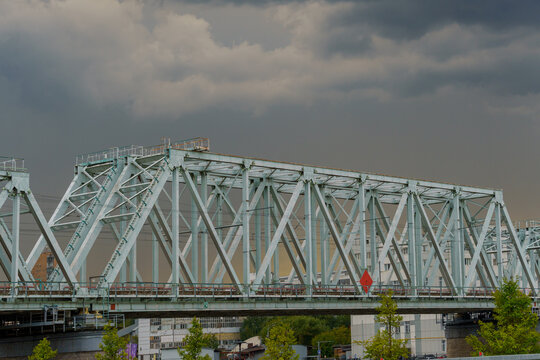 Steel railway bridge truss structure under a dramatic stormy sky background. Urban transport infrastructure network featuring metal construction span over city buildings and green trees.
