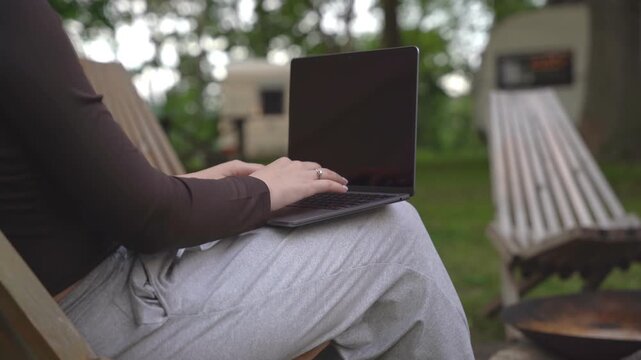 Person typing on laptop while seated outdoors on wooden chair. Background shows green lawn, fire pit, and camper. Scene conveys calm, focused, and relaxed work atmosphere. Ideal for remote work