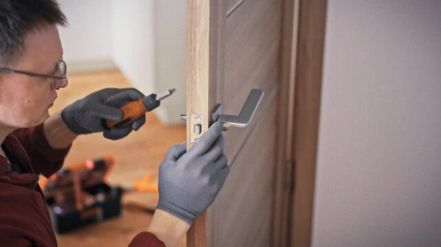 A specialist in work gloves fixes a door handle on a grey brown wooden interior door with an orange screwdriver and orange toolbox visible nearby. Concept of home door maintenance and repair
