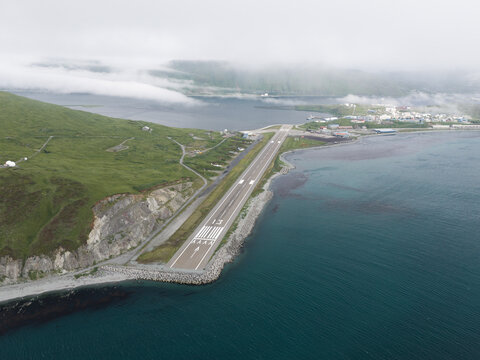 Aerial view of the Unalaska Airport runway situated along the rocky coastline with green hills and calm water in Unalaska, Alaska, United States.