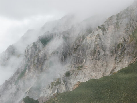 Aerial view of rugged rocky mountainside partially obscured by thick fog with steep cliffs and patches of green vegetation in the Aleutian Islands, Unalaska, Alaska, United States.