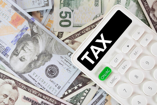 Tax season and accounting concept. White calculator with TAX text on screen lying on a pile of US dollar banknotes. Financial planning, business taxation, and income return preparation.