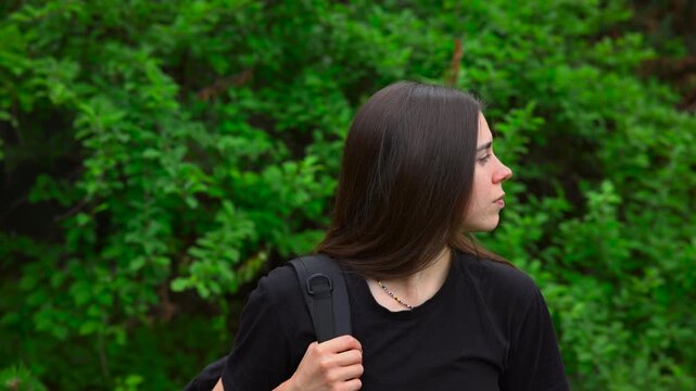 Young woman in side profile holding backpack strap beside bright green foliage, calm outdoor lifestyle moment with soft daylight, shallow depth, and clear space around subject