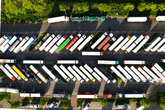 Aerial view of a large truck parking lot filled with numerous semi-trucks and trailers parked in organized rows, surrounded by green trees in Mannheim, Baden-Wurttemberg, Germany.