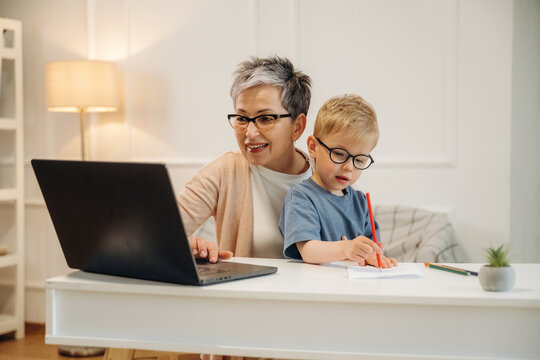 Elderly woman with short gray hair and glasses smiles while using a laptop at a white desk with a young boy wearing glasses drawing beside her in a cozy home environment