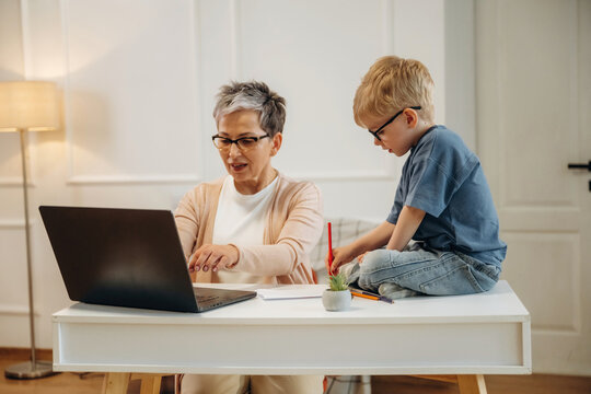 Senior woman with short gray hair using laptop at desk while young boy in glasses sits beside her, engaged in drawing with colored pencils in a cozy home environment