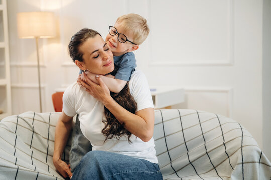 Caucasian woman sitting on a couch with a young boy hugging her from behind, cozy living room setting with soft lighting and modern decor visible in the background
