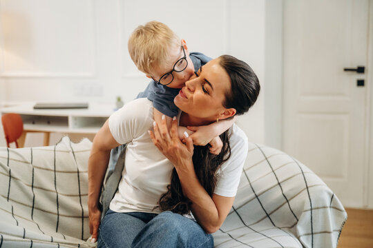 Woman with long dark hair sitting on a couch with a young boy wearing glasses, the boy playfully hugging her from behind in a bright, modern living room