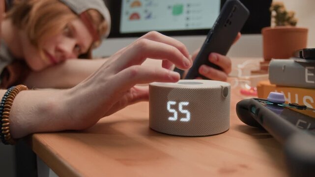 Caucasian male teenager lying on desk, holding smartphone and adjusting smart speaker volume. Footage represents digital distraction and smart home technology usage