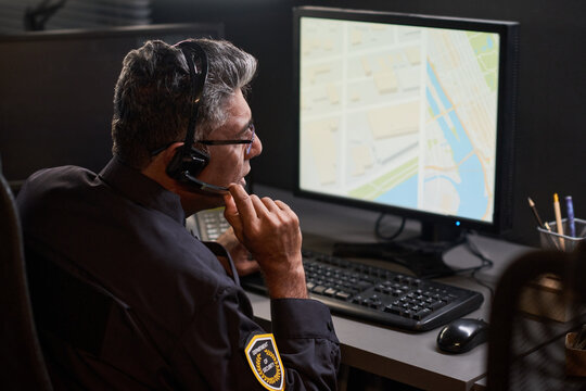 Middle aged security guard monitoring digital map on desktop computer while speaking through headset in control room. Ideal for surveillance, dispatch, safety service marketing