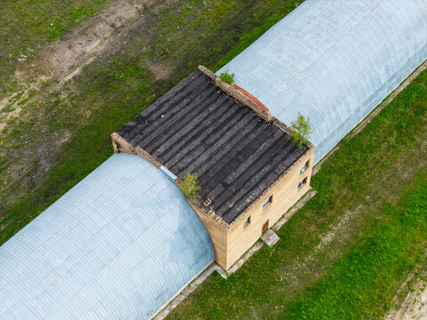 Aerial view of a long arched metal hangar with a central brick building section surrounded by green fields in Kalnciems, Jelgava Municipality, Latvia.