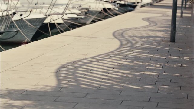 Curved shadows from a marina railing stretch across a paved waterfront promenade beside moored boats in Zadar Croatia.