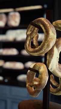 A person admires soft pretzels hanging on a wooden stand in a bakery. Fresh bread loaves fill the shelves in the background. Customers enjoy the smell of baked goods.