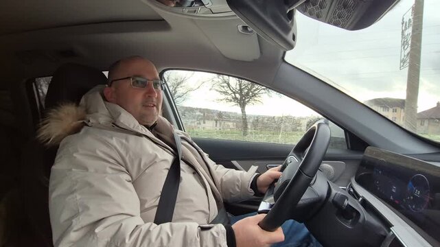 Middle aged man driving luxury car while chatting along road. Interior passenger seat view with steady fixed camera shot.