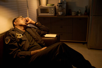 Fototapeta premium Middle aged security guard resting on sofa during break, rubbing forehead in office kitchen, conveying fatigue from long shift. Useful for workplace stress, burnout, night duty campaigns