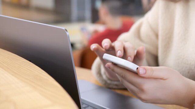 Person uses smartphone beside laptop at wooden desk. Focused on device, hands steady. Background shows blurred indoor space. Ideal for digital work, remote learning, or multitasking themes