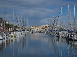 Obraz premium Symmetrical view of sailboats moored in a calm Mediterranean marina with water reflections and waterfront architecture.