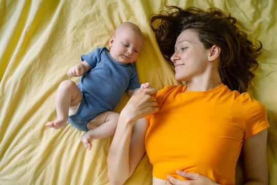 New mother lying on a yellow bed with her baby, connecting through a tender touch, symbolizing early parenthood, bonding, attachment, and the loving development of a family relationship
