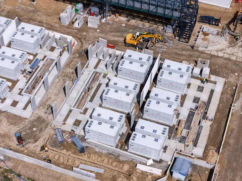 Aerial view of a construction site featuring rows of white industrial battery storage units on concrete pads with an excavator and workers in Poland.