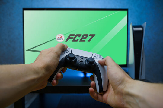 Hands holding PlayStation 5 DualSense controller in front of TV screen showing EA Sports FC 27 game, console gaming and football video game concept.
