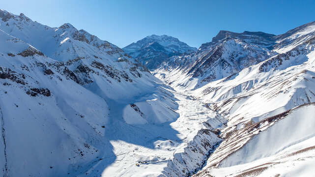 Aerial view of the snow-covered Aconcagua mountain range with deep valleys and rugged peaks under a clear blue sky in Las Heras Department, Mendoza Province, Argentina.