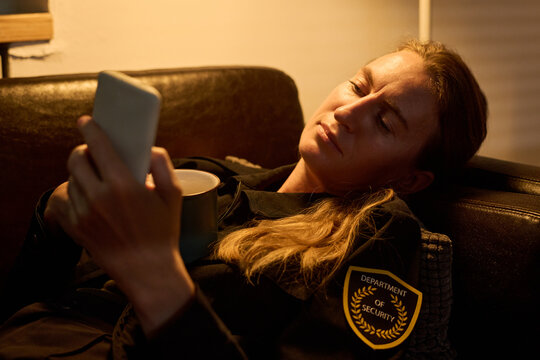 Caucasian woman resting on sofa while checking smartphone and holding mug, suggesting off duty security work. Useful for burnout, shift fatigue, mobile communication themes