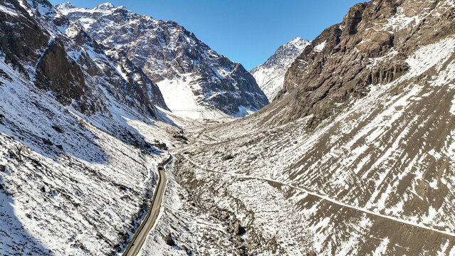 Aerial view of the Andes mountains featuring snow-covered peaks, a winding mountain road, and rugged rocky slopes under a clear blue sky in Portillo, Valparaiso, Chile.