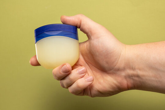 Hand holding a small jar of petroleum jelly against a muted green background