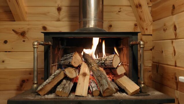 Cozy 4K residential interior scene inside an alpine timber cabin showcasing a heavy cast iron wood burning stove with bright orange flames roaring behind the glass door supplemented by a neat stack