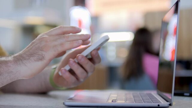 Hand uses smartphone beside laptop. Fingers scroll actively on screen. Background shows blurred cafe space. Person appears focused and engaged. Suitable for tech, communication, or multitasking themes