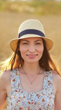 Charming young woman wearing a straw hat and floral dress smiling playfully at the camera while standing in a golden wheat field at sunset, with beautiful backlight highlighting her hair