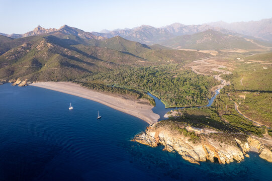 Aerial view of the Fango river mouth meeting the Mediterranean Sea at the beach, surrounded by lush green mountains and rocky cliffs in Galeria, Corsica, France.