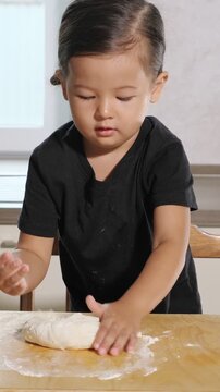 Adorable toddler with a serious expression carefully kneading fresh dough on a wooden board in the kitchen, learning how to bake bread and developing fine motor skills through a fun activity