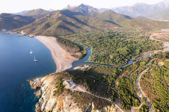 Aerial view of the Fango river mouth meeting the Mediterranean Sea, with a sandy beach, sailboats, lush forests, and rugged mountains in Galeria, Corsica, France.