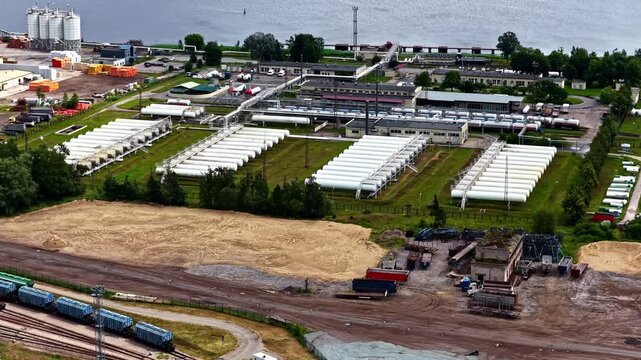 Riga, Latvia - 17 July 2025: Aerial view of industrial facility with rows of white storage tanks and railway tracks near the river.
