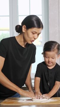 Young mother teaching her cute little girl how to knead dough on a wooden board while cooking together in the kitchen, creating a special moment of learning and bonding