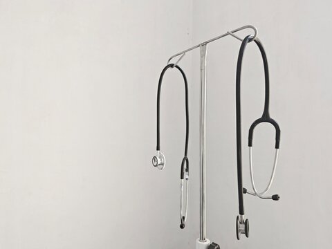 Two stethoscopes hanging on a medical equipment stand in a clean examination room, symbolizing healthcare, clinical diagnosis, and professional medical services with copy space