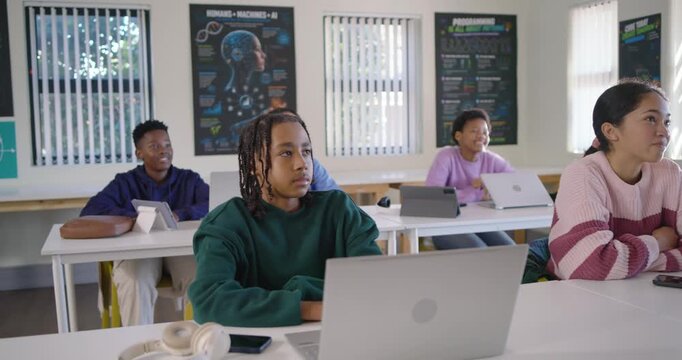 Diverse teens at laptops in classroom watching speaker reacting smiling at male in green sweater