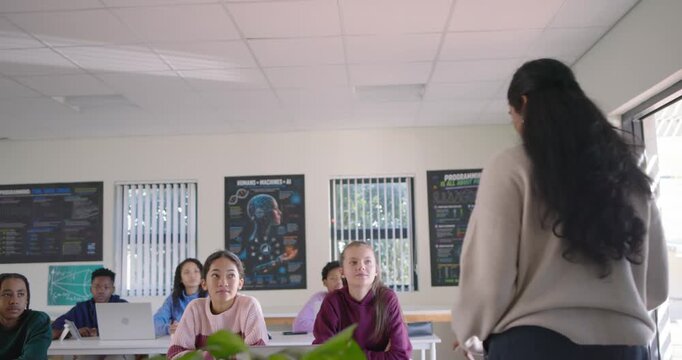 Indian female instructor walking ahead after teens looking up, leaning over laptop in class to help